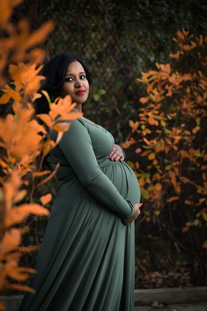 Expectant mother in a green dress posing elegantly among autumn leaves in a serene outdoor setting.